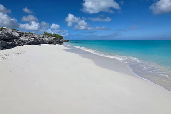 White sand beach with turquoise water, rocky outcrop with vegetation, and blue sky with white clouds