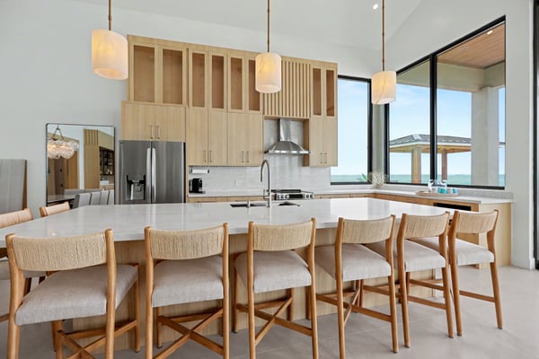 Modern kitchen and dining area with light wood cabinetry, stainless steel appliances, white quartz island, wooden chairs, and ocean view through large windows.